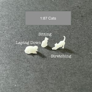 May include: Four white plastic miniature cats in different poses: sitting, laying down, and stretching. The text "1:87 Cats" is above the cats. The text "Sitting", "Laying Down", and "Stretching" are below the cats.