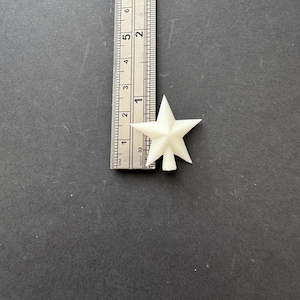 May include: A small, off-white star-shaped object is positioned next to a ruler. The ruler shows measurements in inches. The star appears to be approximately 2.5 cm in diameter. The background is a solid, dark grey surface.