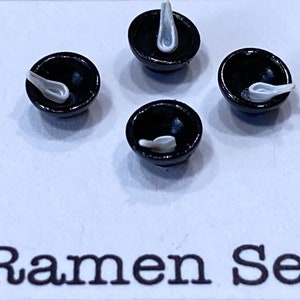 Puede incluir: Un juego de cuatro cuencos de ramen en miniatura negros con cucharas blancas. Los cuencos están dispuestos en un patrón cuadrado sobre una superficie blanca. El texto "Ramen Set" está impreso debajo de los cuencos.