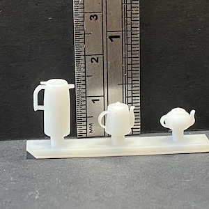 May include: Three white ceramic teapots on a white base. The teapots are different sizes and shapes. The largest teapot has a handle on the side and a spout on the top. The middle teapot has a handle on the side and a spout on the top. The smallest teapot has a handle on the side and a spout on the top.