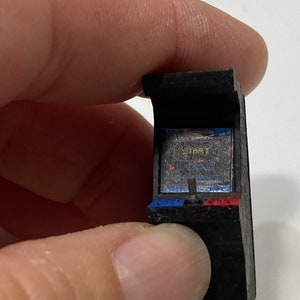 May include: A miniature black arcade cabinet with a blue and red base. The screen displays the word "START" in yellow text.