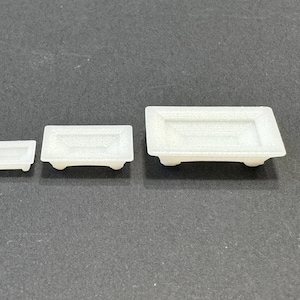 May include: Three white ceramic rectangular trays of varying sizes. The trays have a raised lip around the edge and four small feet on the bottom.