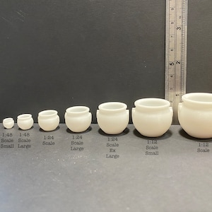 May include: Seven white ceramic pots of varying sizes, arranged from smallest to largest. The pots have a rounded shape with two small handles on each side. The text below the pots indicates the scale of each pot: 1:48 Scale Small, 1:48 Scale Large, 1:24 Scale, 1:24 Scale Large, 1:24 Scale Ex Large, 1:12 Scale Small, 1:12 Scale.