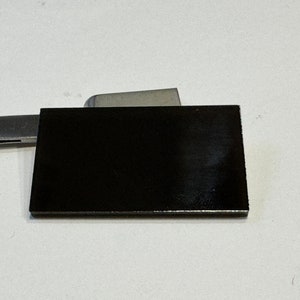 May include: A rectangular, black object with a glossy finish. It rests on a white surface, with a metal tool partially visible in the background. The object's edges are sharp and well-defined.