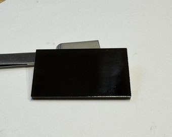 May include: A rectangular, black object with a glossy finish. The object is positioned on a white surface, with a metal tool partially visible in the background. The object's edges appear sharp and well-defined.