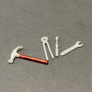 Może przedstawiać: Zestaw miniaturowych narzędzi, w tym młotek z czerwonym uchwytem, szczypce, śrubokręt i klucz. Narzędzia wykonane są ze srebrnego metalu.
