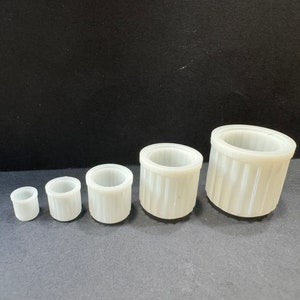 May include: Five white plastic containers of varying sizes. The containers are cylindrical and have a smooth, slightly textured surface.