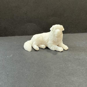 Puede incluir: Figura de cerámica blanca de un perro acostado. El perro tiene una cola esponjosa y mira hacia la derecha.