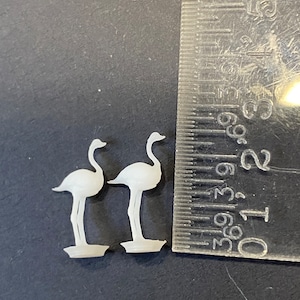 May include: Two white plastic flamingo figurines standing on a black surface. The figurines are about 2 inches tall and are posed with their necks bent.