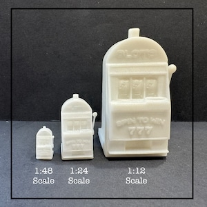 May include: Three white slot machine models of varying sizes, labeled 1:48, 1:24, and 1:12 scale. The largest model has the text "SLOTS" and "SPIN TO WIN 777". Each model features three reels and a lever.