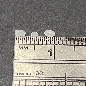 May include: Four small, white, round, plastic objects are lined up on a ruler. The ruler is marked in millimeters and inches. The objects are about 1 centimeter in diameter.