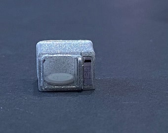 Könnte beinhalten: Ein silberfarbener Miniatur-Mikrowellenherd mit einer quadratischen Glastür, einem kleinen runden Drehteller im Inneren und einem vertikalen Bedienfeld auf der rechten Seite mit einem dunklen, rechteckigen Displaybereich.