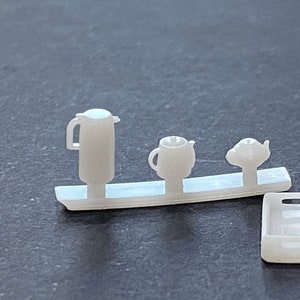 May include: A sprue of miniature white plastic tea party accessories. The sprue includes a tall thermos, two teapots, and a small tray. The objects are all white and appear to be made of plastic. The background is a dark gray surface.
