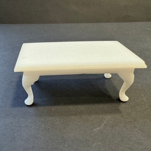 May include: A small, white table with a rectangular top and four curved legs. The table is likely a miniature or dollhouse furniture piece, with a classic design. The table's dimensions appear to be approximately 15 cm long, 10 cm wide, and 8 cm high.