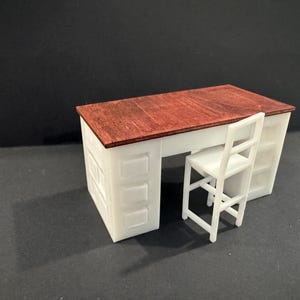 Könnte beinhalten: Ein Miniaturschreibtisch mit einer dunkelrotbraunen Holzmaserungsplatte und einem weißen Unterbau mit geformten Schubladendetails an den Seiten. Ein passender weißer Stuhl mit hoher Rückenlehne und vier Beinen steht vor dem Schreibtisch.