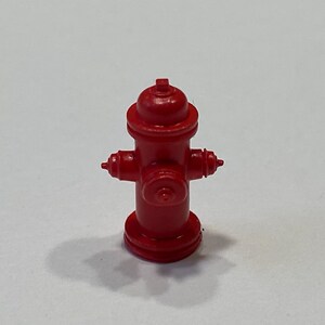 Op de afbeelding: Een kleine, felrode brandkraan. De brandkraan heeft een afgeronde bovenkant, twee zijsproeiers en een cirkelvormige basis. Het is een miniatuurreplica van een standaard brandkraan.