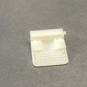 Könnte beinhalten: Ein weißes Plastik-Spielzeug im Miniaturformat einer Schreibmaschine. Die Schreibmaschine hat eine schwarze Tastatur und einen weißen Wagenrücklaufhebel.