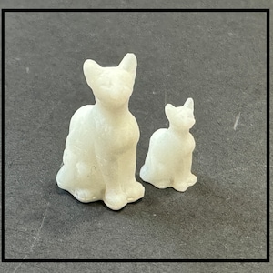 Może przedstawiać: Dwie białe ceramiczne figurki kotów. Większy kot siedzi z łapami schowanymi pod ciałem. Mniejszy kot siedzi z łapami wyciągniętymi do przodu.