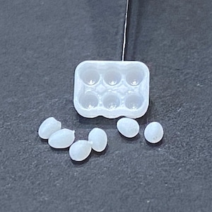 May include: A small, translucent plastic egg carton with six circular indentations, next to several small, white, oval-shaped objects resembling eggs. The objects are scattered on a dark gray surface. The image is well-lit, highlighting the details of the objects.