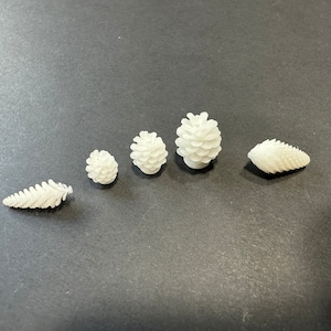 May include: Five white pine cone ornaments of varying sizes. The ornaments are arranged in a row on a dark gray surface.
