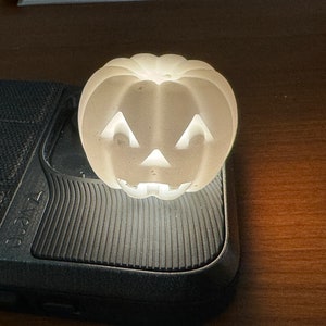 May include: A white ceramic pumpkin with a carved jack-o'-lantern face. The pumpkin is lit from inside, creating a warm glow.