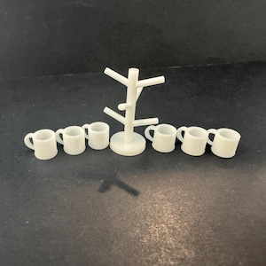 May include: A white miniature mug tree stand with a central pole and six angled branches, accompanied by six small white mugs. The set is arranged with the stand in the center and three mugs placed on either side of the base on a flat surface.