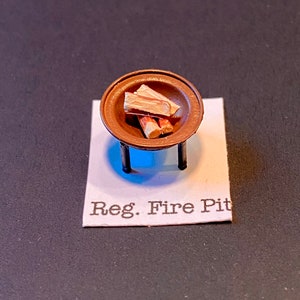 Puede incluir: Un hogar de fuego en miniatura con una base de metal marrón y un cuenco redondo. El cuenco contiene tres pequeños trozos de madera, que se asemejan a una fogata. El hogar de fuego está etiquetado como "Reg. Fire Pit".