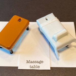 Puede incluir: Dos mesas de masaje en miniatura, una marrón y otra blanca, con el texto "Massage table" en una tarjeta blanca.
