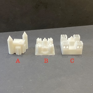 Puede incluir: Se muestran tres pequeños modelos blancos de castillos en miniatura. Cada castillo tiene un diseño único, con torres, muros y detalles arquitectónicos. Los modelos están etiquetados A, B y C, sobre un fondo oscuro.