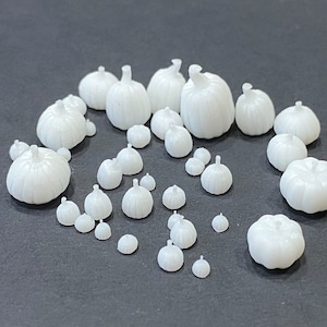 May include: A collection of white ceramic pumpkins of various sizes, ranging from small to large. The pumpkins are arranged in a cluster on a gray surface.