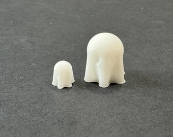 May include: Two small, white, ghost-shaped figurines. The larger ghost has a rounded top and a draped, flowing body. The smaller ghost has a similar design, but is significantly smaller. Both are set against a dark gray background.