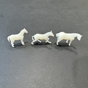 May include: Three small, off-white horse figurines in various poses. One stands, one is mid-stride, and the third is grazing. The figurines are set against a dark background.
