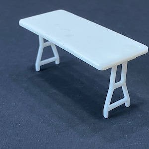 May include: A small, white rectangular table with a flat top and A-frame legs. The table is made of a solid, opaque material and is designed for miniature settings. The table measures approximately 4 inches long.