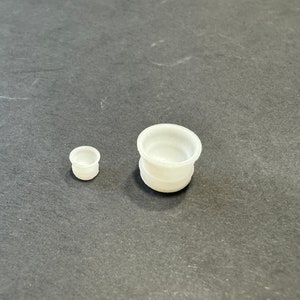 Puede incluir: Dos objetos cilíndricos blancos pequeños con una parte superior redondeada y un borde ligeramente ensanchado. Uno es más pequeño que el otro. Los objetos están hechos de un material translúcido y se encuentran sobre una superficie gris oscuro.