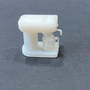 May include: A small, off-white coffee maker model. The miniature appliance features a rectangular base, a cylindrical body, and a carafe with a handle. The coffee maker has a spout and a top section with a rectangular design.