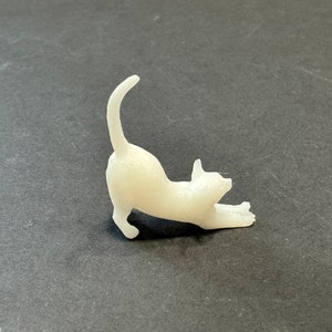 May include: A small, off-white cat figurine in a stretching pose. The cat is arched, with its front paws extended and tail curved upwards. The figurine is on a dark gray surface.