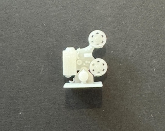Könnte beinhalten: Ein kleines, cremefarbenes Modell eines Vintage-Filmprojektors. Der Projektor hat zwei Filmrollen, eine Linse und einen rechteckigen Körper. Das Modell besteht aus einem festen Material und steht auf einem kleinen Sockel. Das Design ist detailliert.