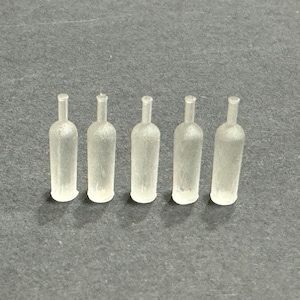 May include: Five small, clear, glass wine bottles are arranged in a row. Each bottle has a long neck and a rounded body. The bottles are made of a translucent material, and they are sitting on a gray surface.