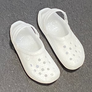 Puede incluir: Un par de zuecos de plástico blanco con una correa en la parte superior. Los zuecos tienen una superficie texturizada y pequeños agujeros por todas partes.