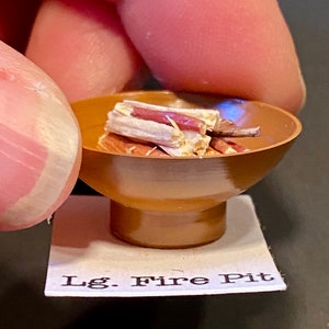 Puede incluir: Un hogar de fuego de cerámica marrón en miniatura con una base y una pila de pequeños troncos de madera en su interior. El hogar de fuego está etiquetado como "Lg. Fire Pit".