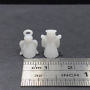 Puede incluir: Dos figuritas de ángel de plástico blanco, una con los brazos extendidos y la otra con alas, se muestran junto a una regla que mide 2,5 cm.