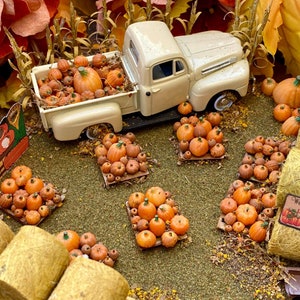 May include: A miniature white pickup truck with a bed full of orange pumpkins. The truck is parked in a miniature pumpkin patch with hay bales and a sign that reads "Welcome to the Pumpkin Patch".