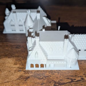 May include: Three white miniature house models with detailed architectural features, including chimneys, gabled roofs, and windows. The houses are arranged on a wooden surface, showcasing their intricate designs and textures. The models are likely for a hobby or craft project.