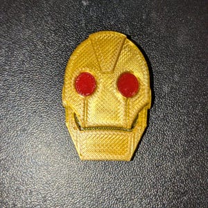 May include: A gold-colored 3D printed robot head with red eyes. The robot head is a small, decorative object.