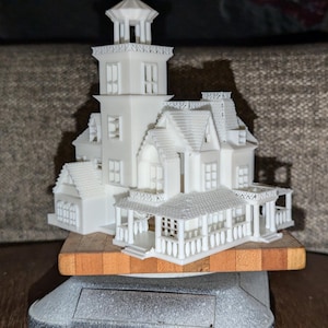 May include: A white 3D printed model of a haunted house with a tower, multiple windows, and a porch. The model is sitting on a wooden base.