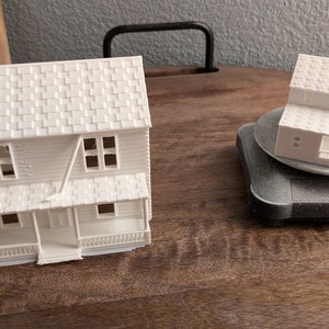 May include: Two white 3D printed model houses. The larger house has a porch and a gabled roof. The smaller house has a simple roof and is on a rotating base.