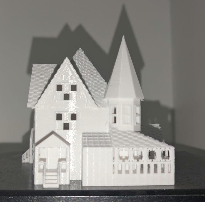 Peut inclure: Mod&egrave;le imprim&eacute; en 3D d'une maison blanche avec des d&eacute;tails complexes. La maison pr&eacute;sente un toit &agrave; pignon, une tour avec un sommet pointu et un porche. Le mod&egrave;le est pos&eacute; sur un fond uni, avec des ombres cr&eacute;ant de la profondeur.