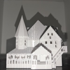 Peut inclure: Une maquette blanche d'une maison miniature avec un design d&eacute;taill&eacute;, comprenant une tour, plusieurs toits &agrave; pignons et un porche. Le mod&egrave;le a une cl&ocirc;ture et des escaliers. La maison est plac&eacute;e sur un fond sombre, avec des ombres projet&eacute;es sur le mur.