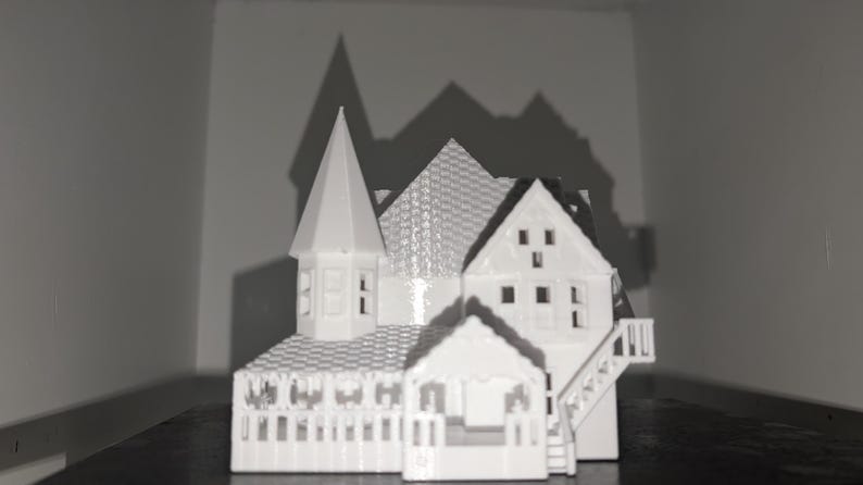 Peut inclure: Une maquette blanche d'une maison miniature avec un design d&eacute;taill&eacute;, comprenant une tour, plusieurs toits &agrave; pignons et un porche. Le mod&egrave;le a une cl&ocirc;ture et des escaliers. La maison est plac&eacute;e sur un fond sombre, avec des ombres projet&eacute;es sur le mur.