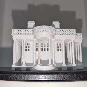 May include: A white miniature model of a classical building with columns, windows, and a detailed facade. The model has a symmetrical design and is likely a decorative item or architectural representation.